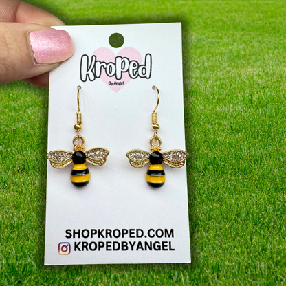 Bee-shaped earrings on a Kroped by Angel card against a green grass background
